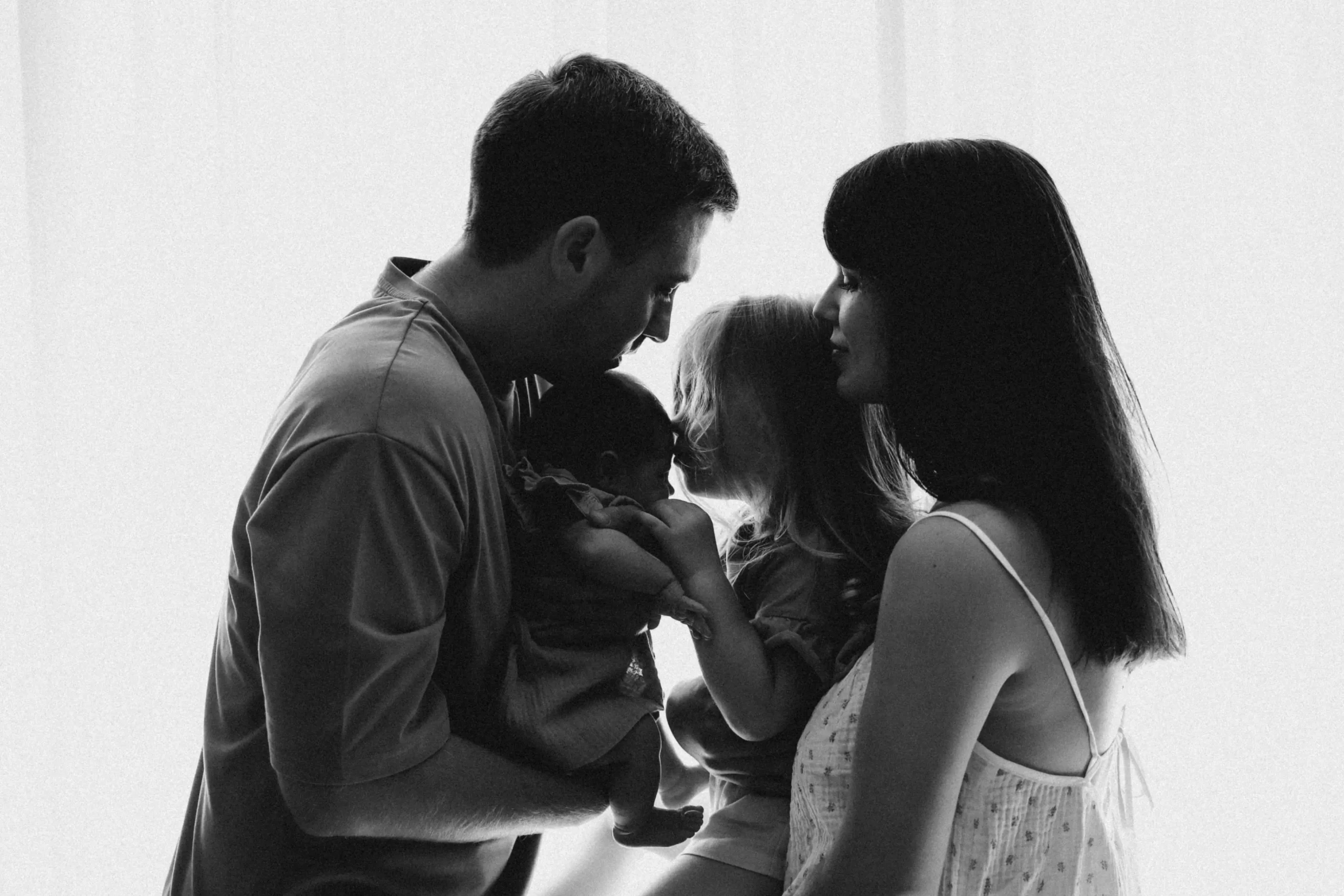 A man and woman stand facing each other, each holding a child, with soft backlighting creating a silhouette effect.