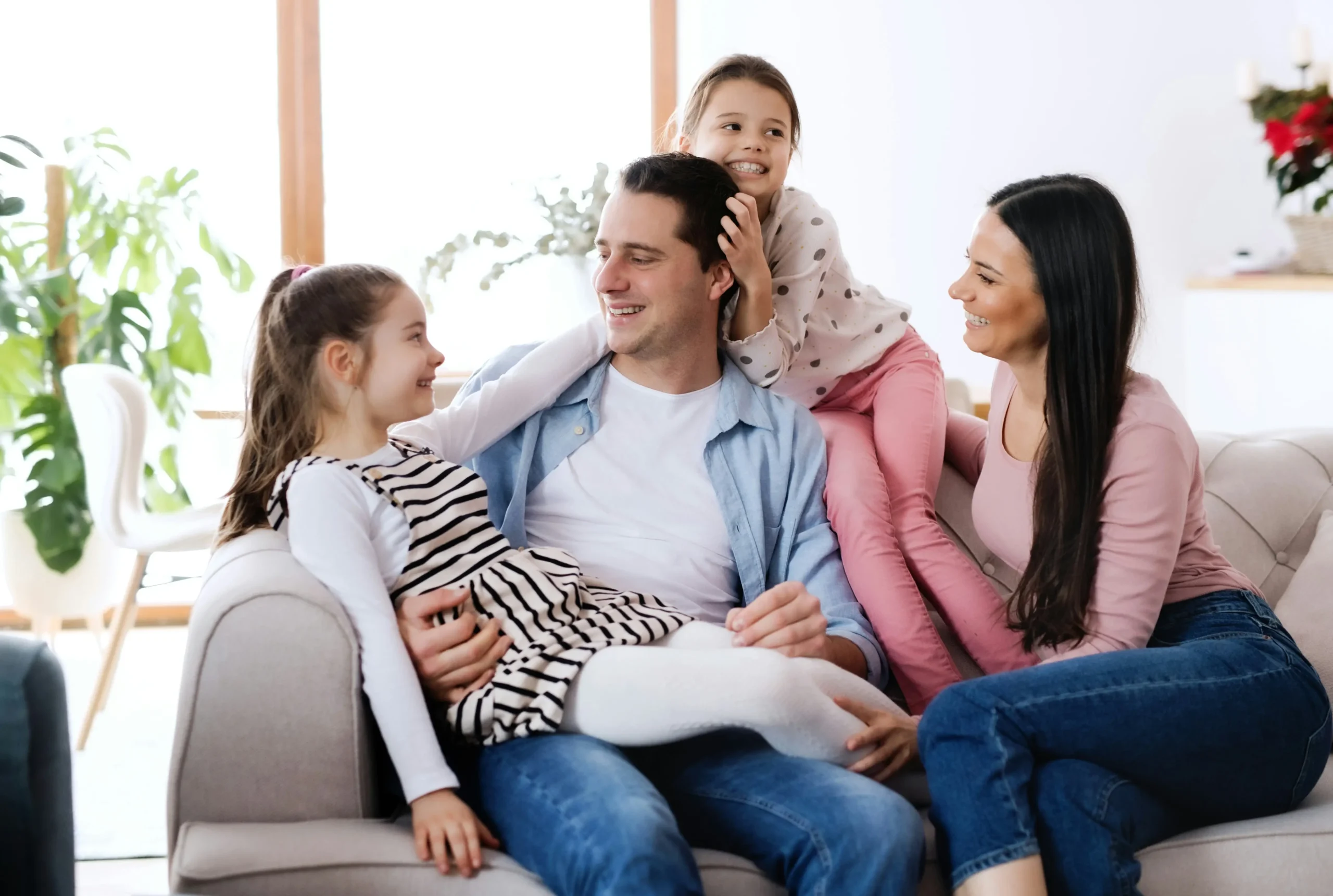 A family of four sits together on a sofa, smiling and interacting in a bright living room with plants in the background.