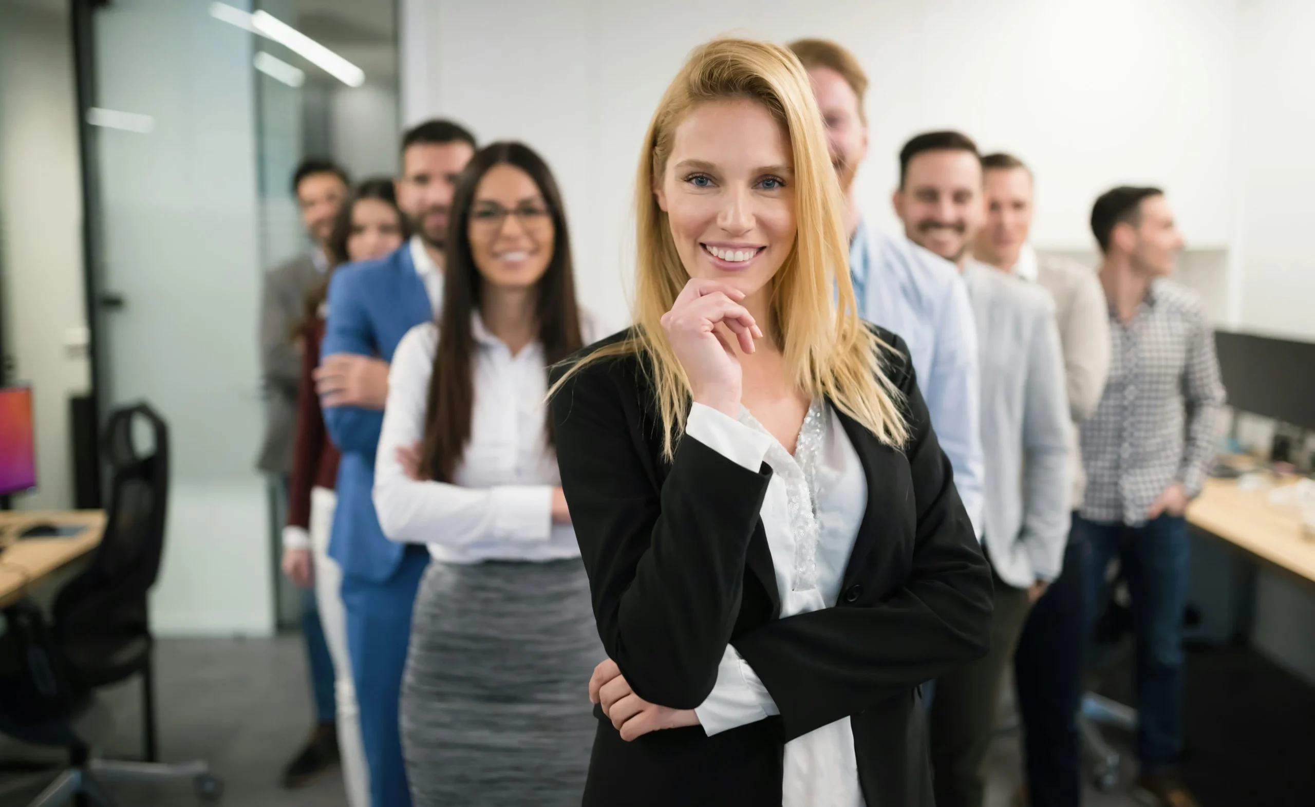 A group of professionals in business attire standing in an office, with a woman in a black blazer smiling at the front.