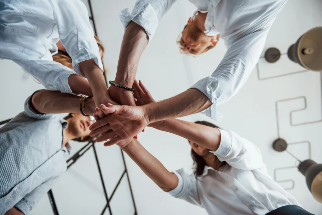 A group of people standing in a circle join their hands together in the centre, viewed from below.