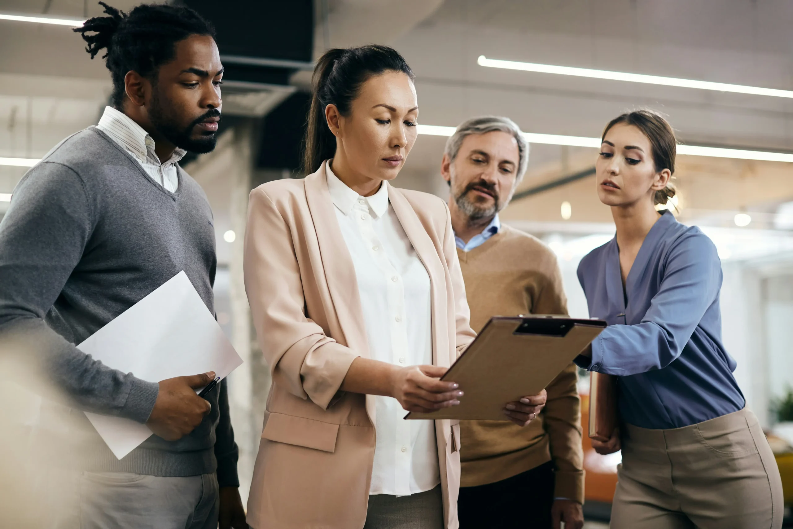 Four people in business attire stand together in an office, looking at a clipboard held by the woman in the centre.
