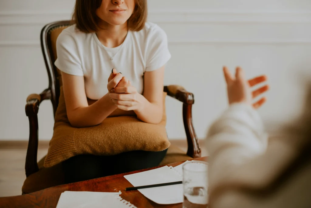 A woman sits in a chair holding a cushion on her lap, listening to another person whose hand is visible in the foreground. Papers and a glass of water are on the table.