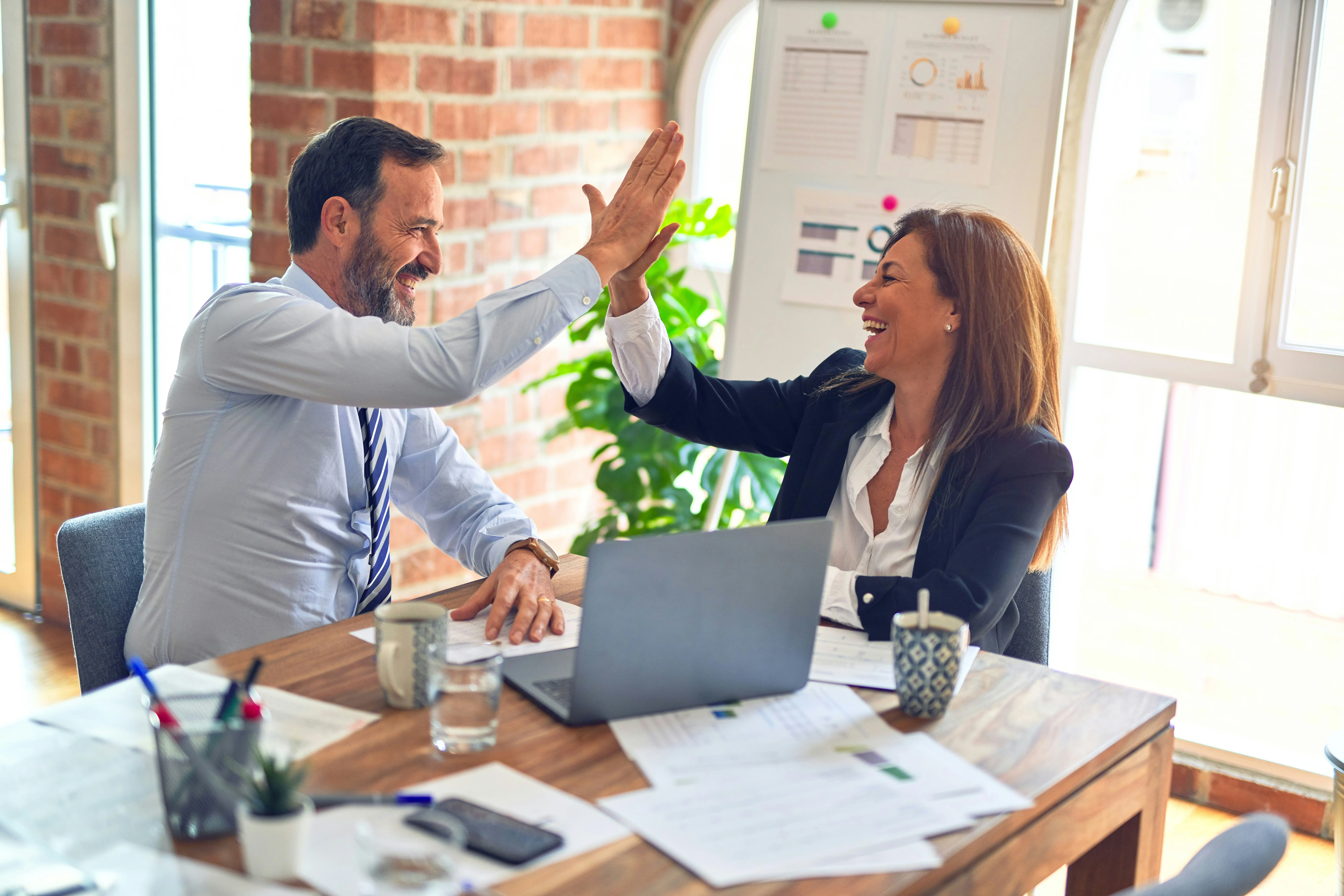 Two colleagues in business attire sit at a desk, smiling and giving each other a high five, with documents and a laptop on the table.