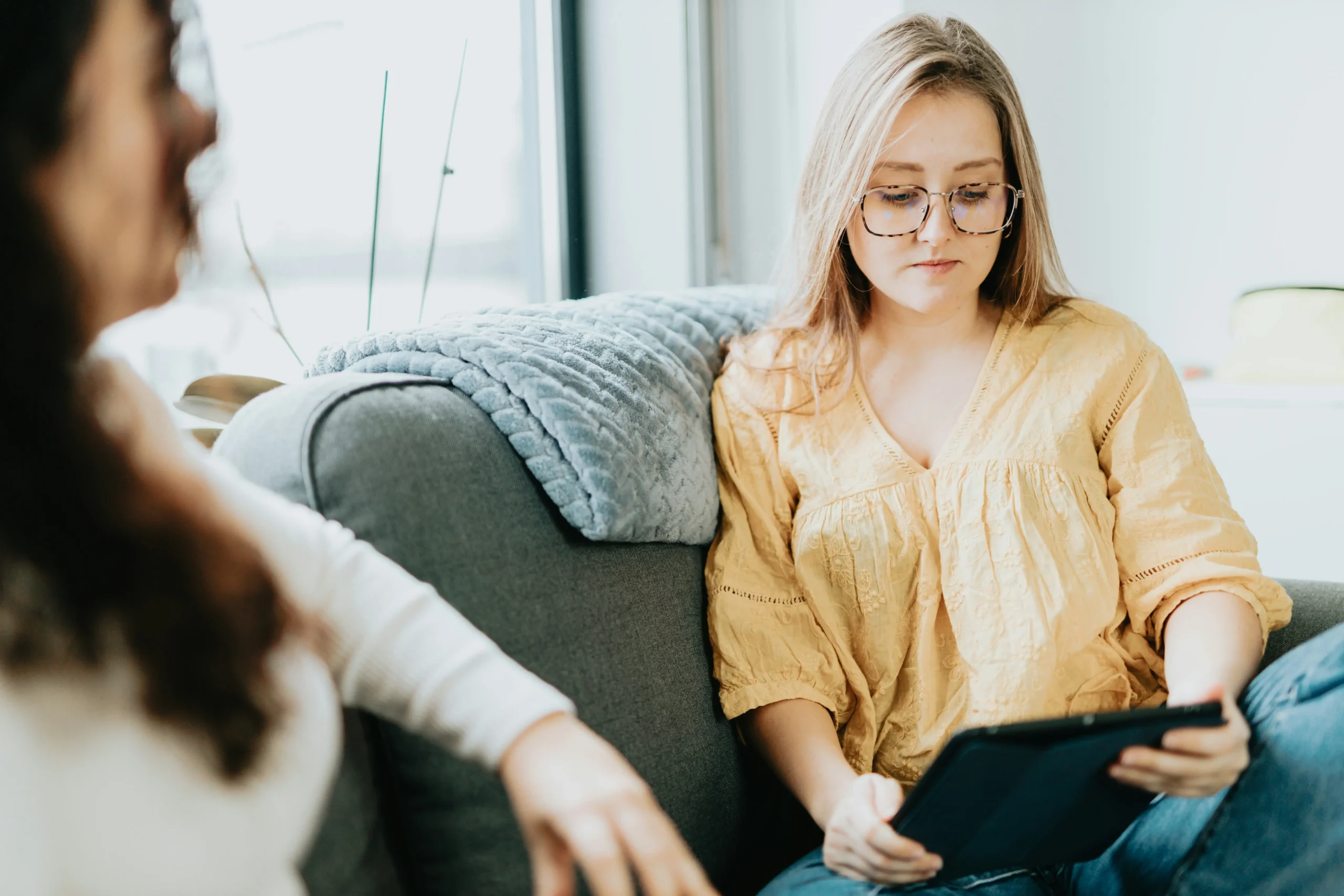 Two people sit on a grey sofa; one woman in a yellow blouse and glasses looks down at a tablet whilst the other person is partially visible on the left.