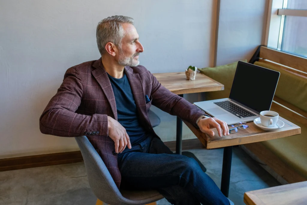 A middle-aged man sits at a table in a café, looking out the window with a laptop, glasses, and a cup of coffee in front of him.