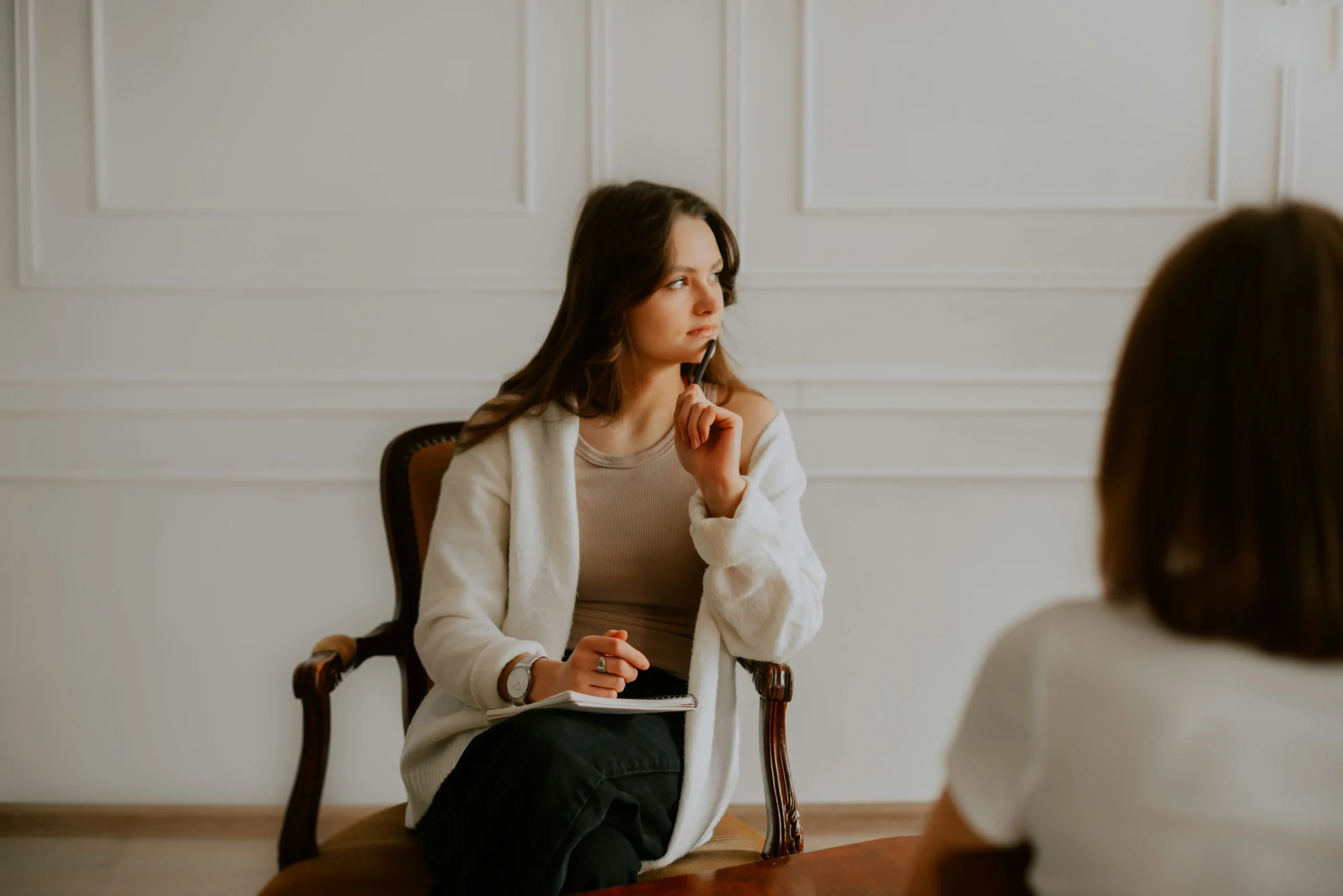 A woman sits on a chair holding a pen and notebook, appearing thoughtful, whilst facing another person in a bright room with white panelled walls.