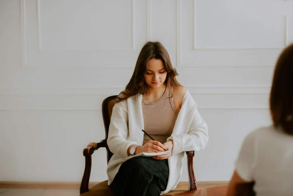 A woman sits in a chair taking notes on a notepad, facing another person whose back is partially visible in the foreground.